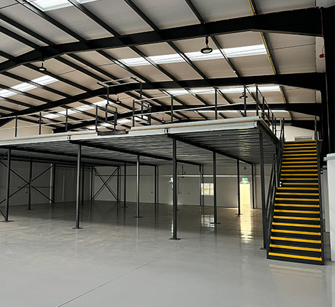 a spacious industrial warehouse featuring a mezzanine level supported by metal beams and a staircase with yellow stripes ideal for storing 13 items or more